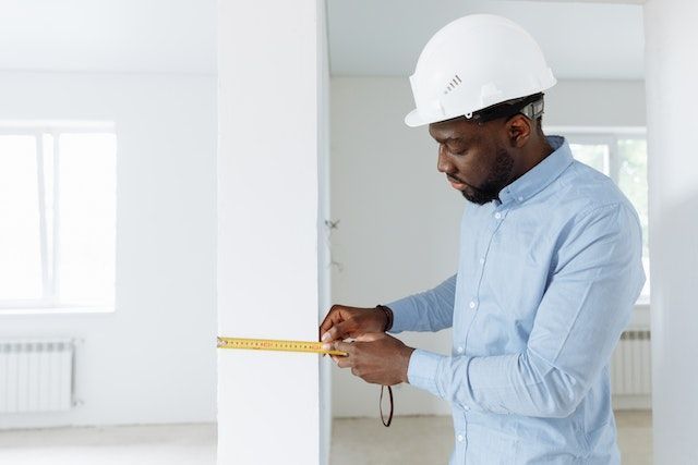 contractor in a blue shirt and white hardhat using a measuring tap