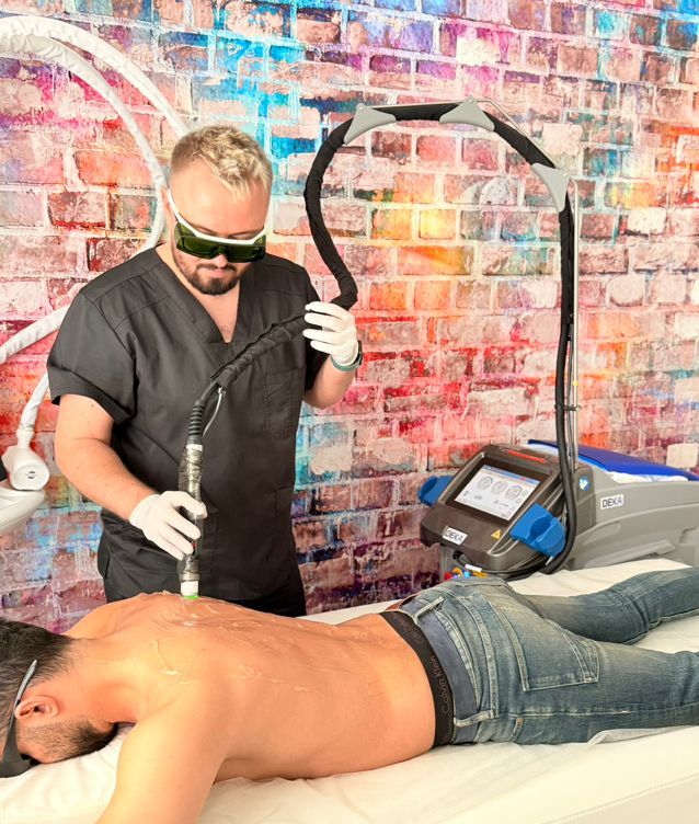 Alex führt eine Rückenbehandlung mit dem Alexandrit-Laser DEKA Motus AX durch. Beide tragen Schutzbrillen; Handschuhe und Kontaktgel sorgen für Sicherheit und Komfort – Laser-Haarentfernung im Studio in Haidhausen, nahe Ostbahnhof. Laser-Haarentfernung am Rücken mit Alexandrit-Laser (Motus AX) im Yuliia Cheporska Studio, München-Haidhausen