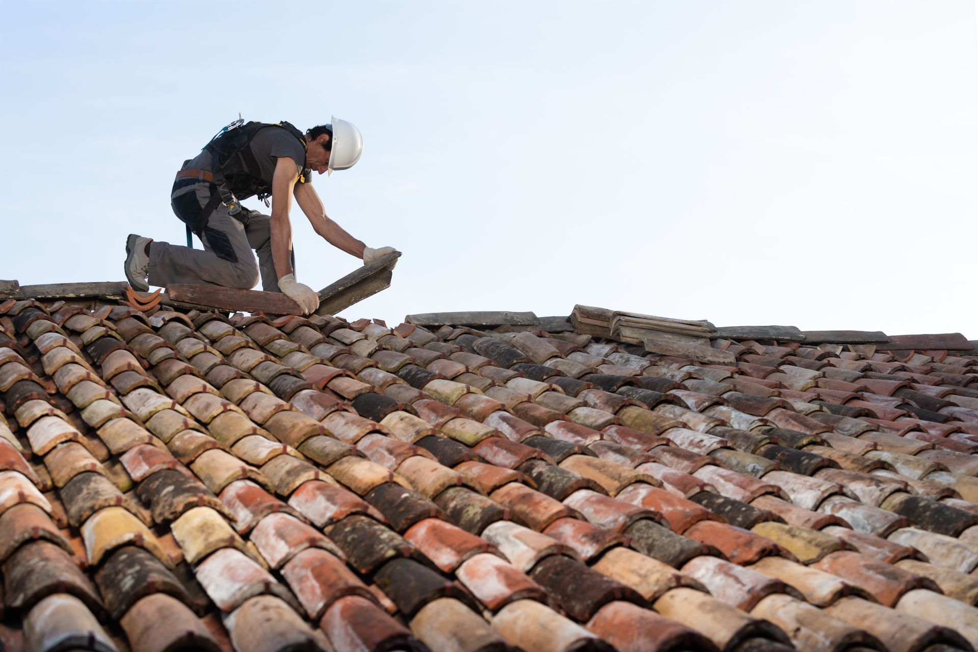 A roofer with a helmet and harness carefully installs clay tiles on a sloped roof.
