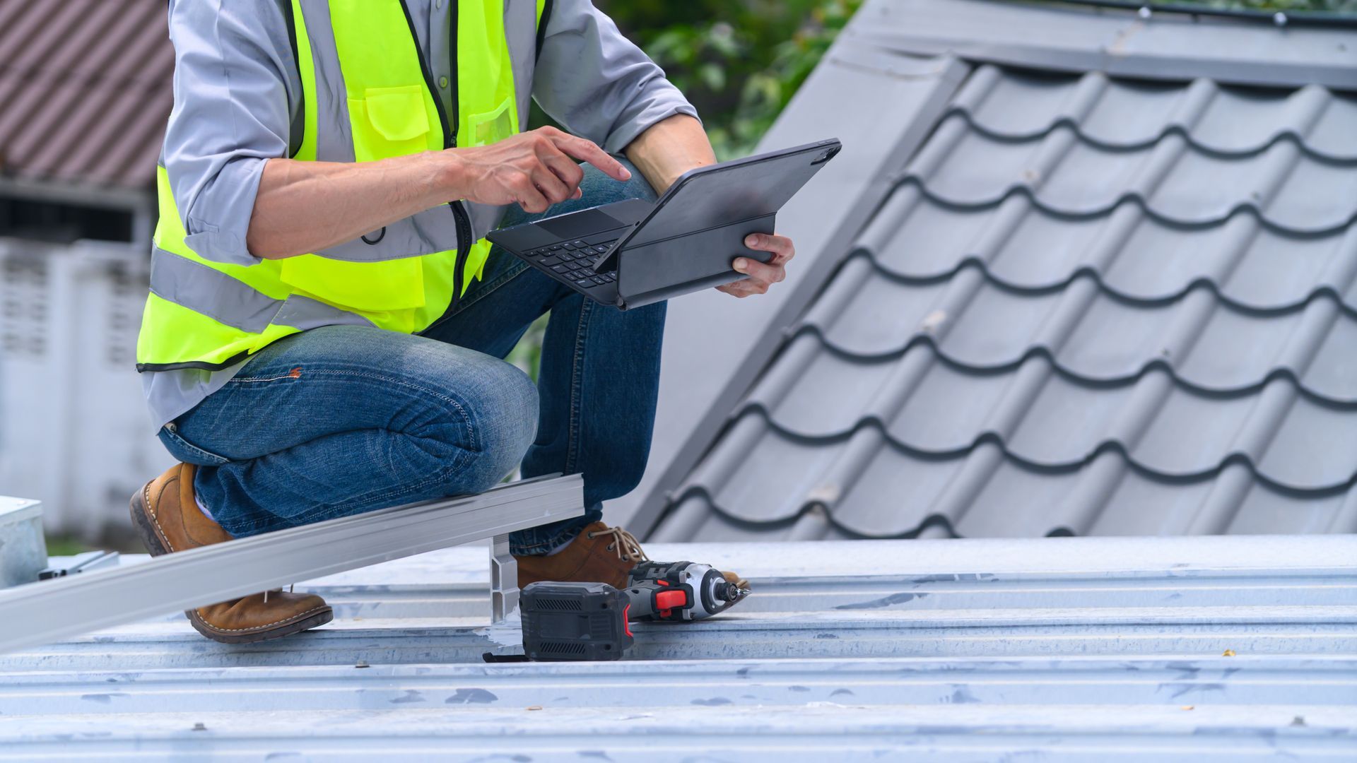 Worker wearing a safety vest and jeans, kneeling on a metal roof while using a laptop
