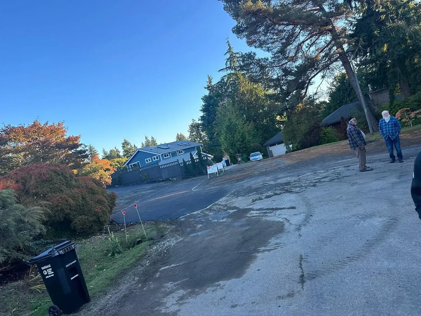A sloped road with a large building, trees, and two people standing. A trash can is in the foreground.