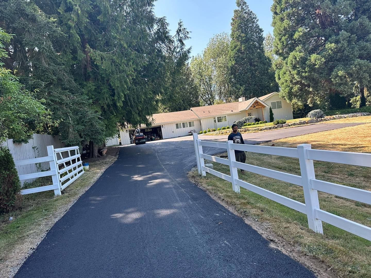 Black asphalt driveway leading to a white house, white fence, and person walking. Green trees and grass are surrounding.