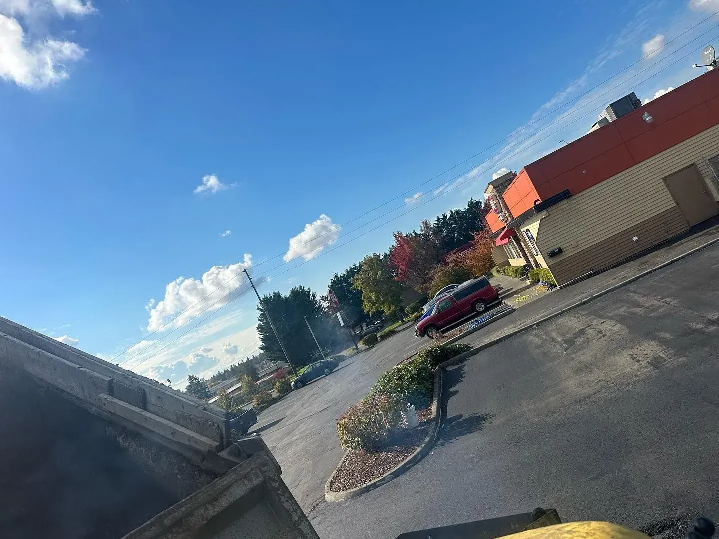 Sunny parking lot with red car, building, blue sky and clouds.