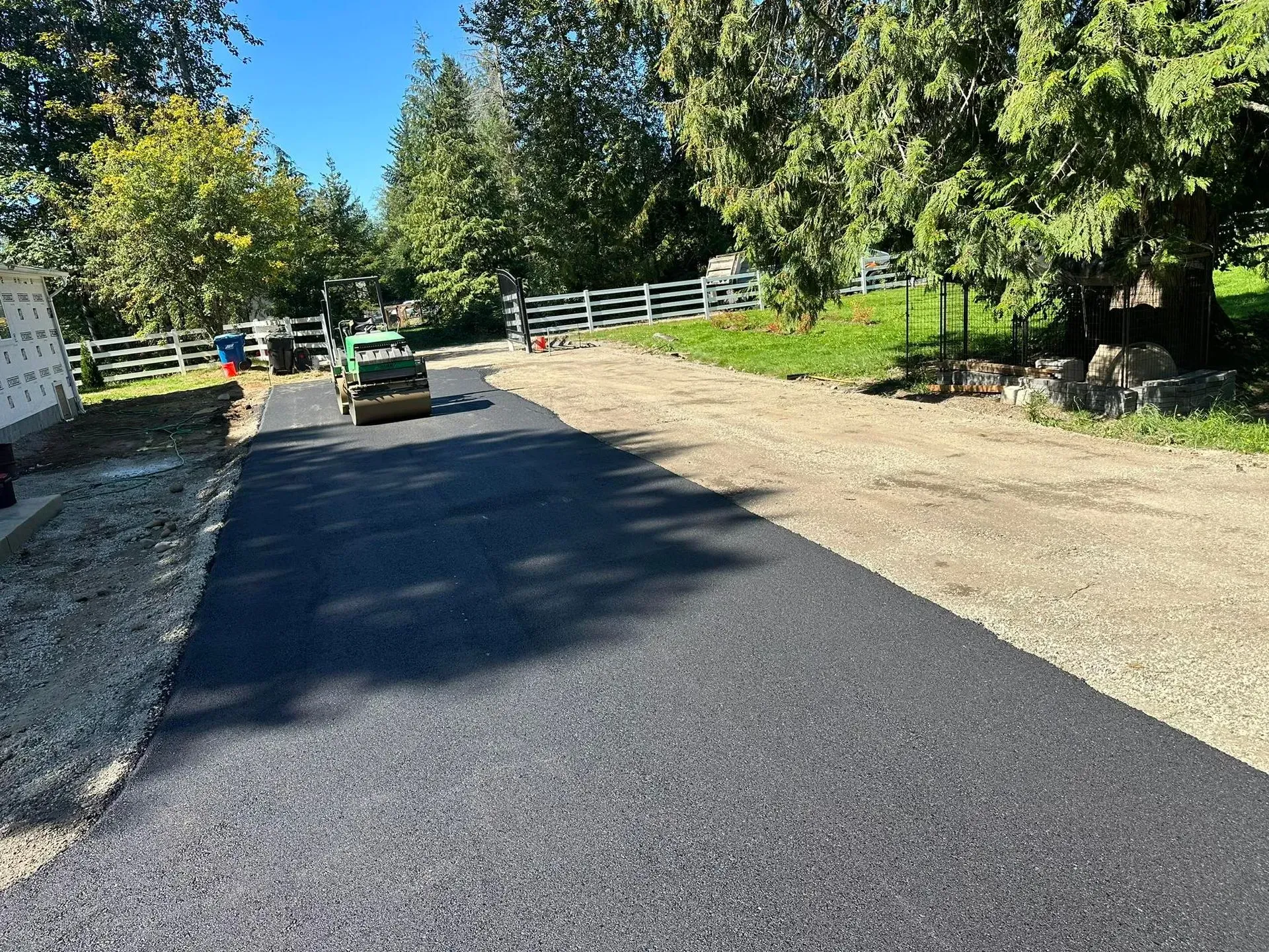 Asphalt driveway being rolled by a small green compactor. Gravel shoulder on the right.