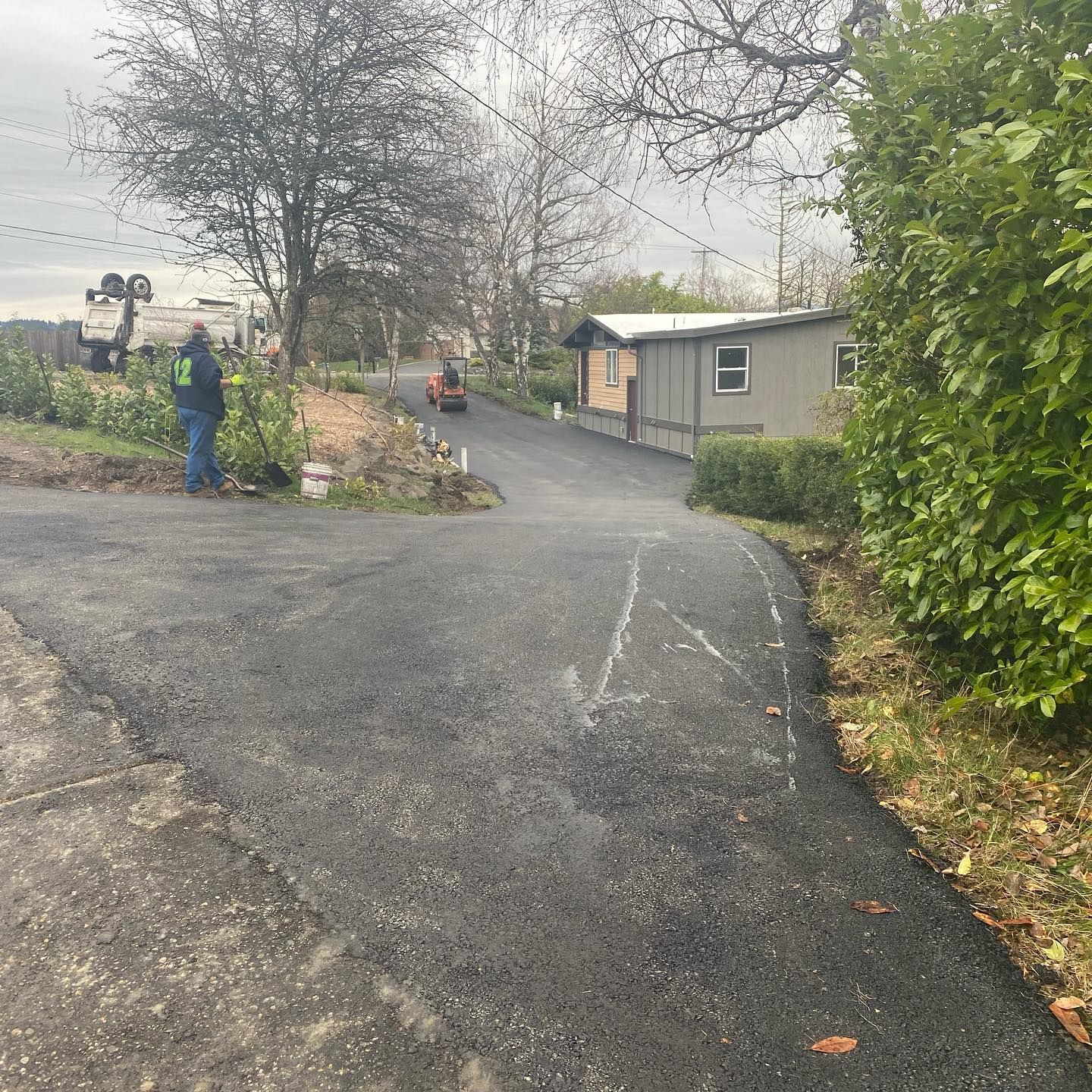 Asphalt driveway with construction equipment and a small building. A person in green jacket stands near a vehicle.