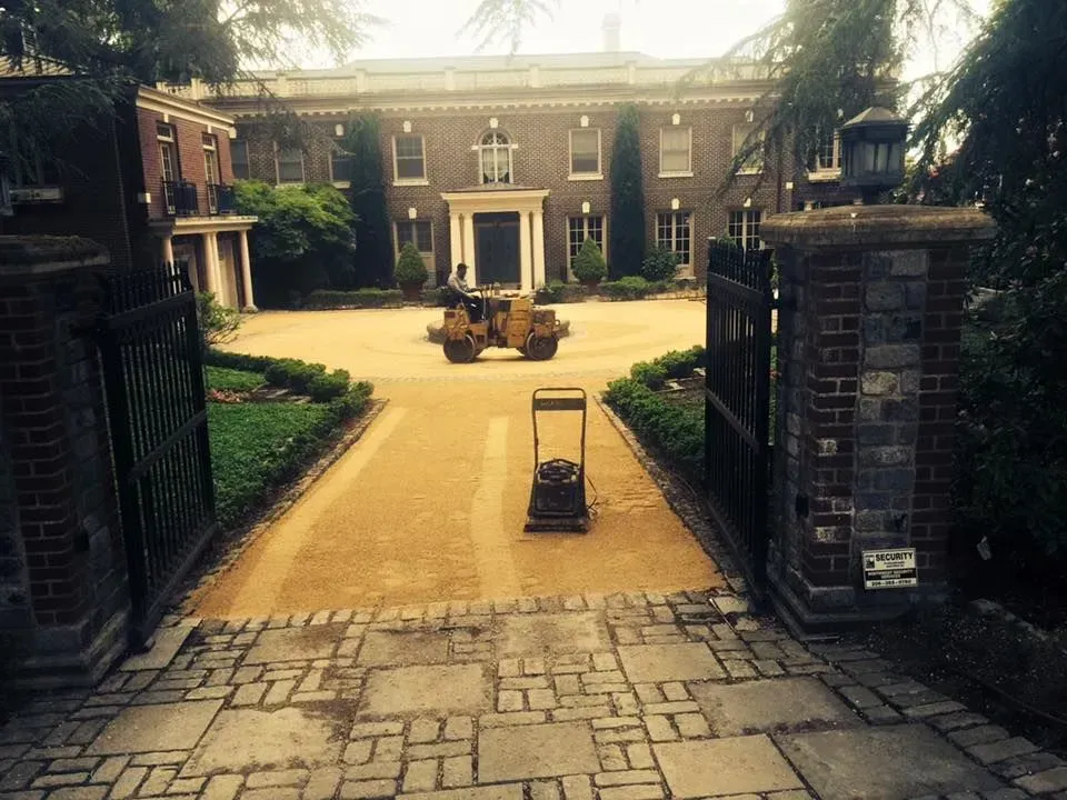 Driveway paving in progress at a large brick house. A roller and compactor are on the new surface.