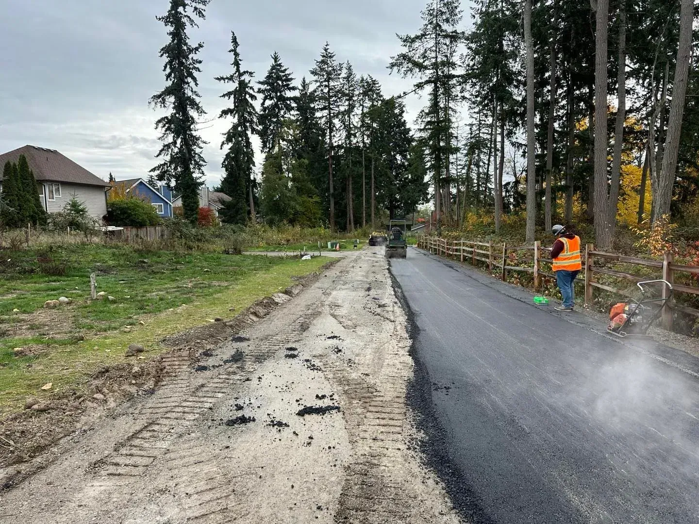 Road construction: A paved road with a worker using a saw, and a muddy shoulder next to a forested area.
