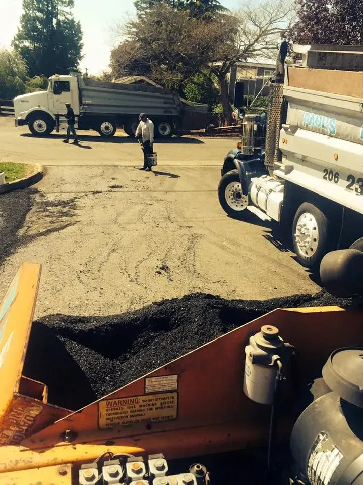 Paving crew with two dump trucks and asphalt laying machine resurfacing a driveway on a sunny day.