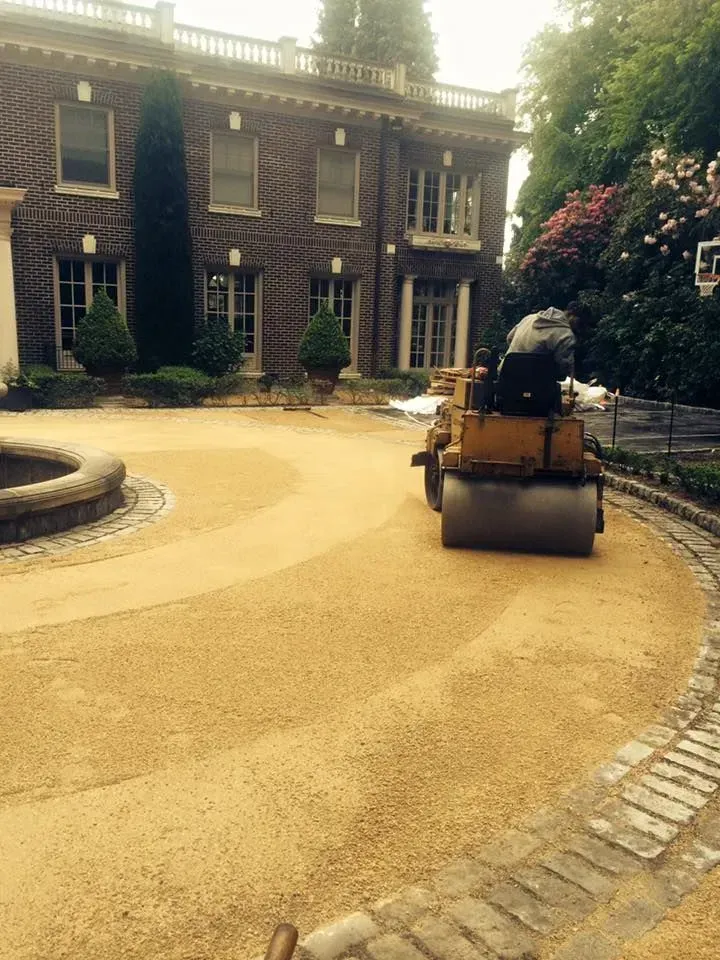 Road roller compacting gravel driveway in front of a large brick house.