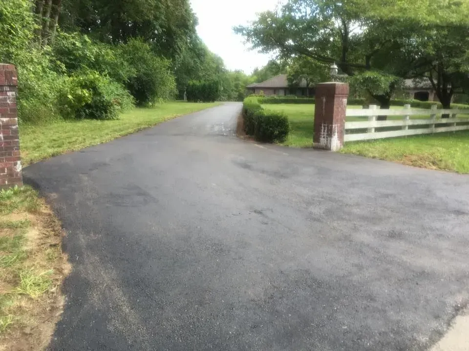 Paved driveway leading toward a house with white fence, brick pillars, and greenery on a cloudy day.