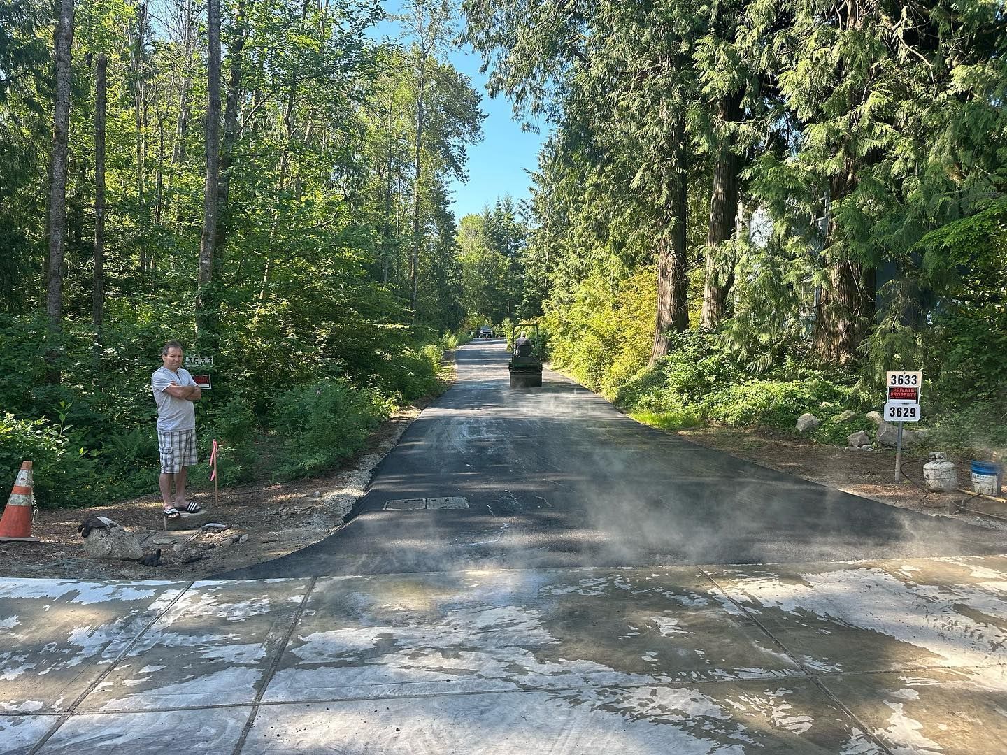 Man watches road paving on a tree-lined street, a construction vehicle in the distance.