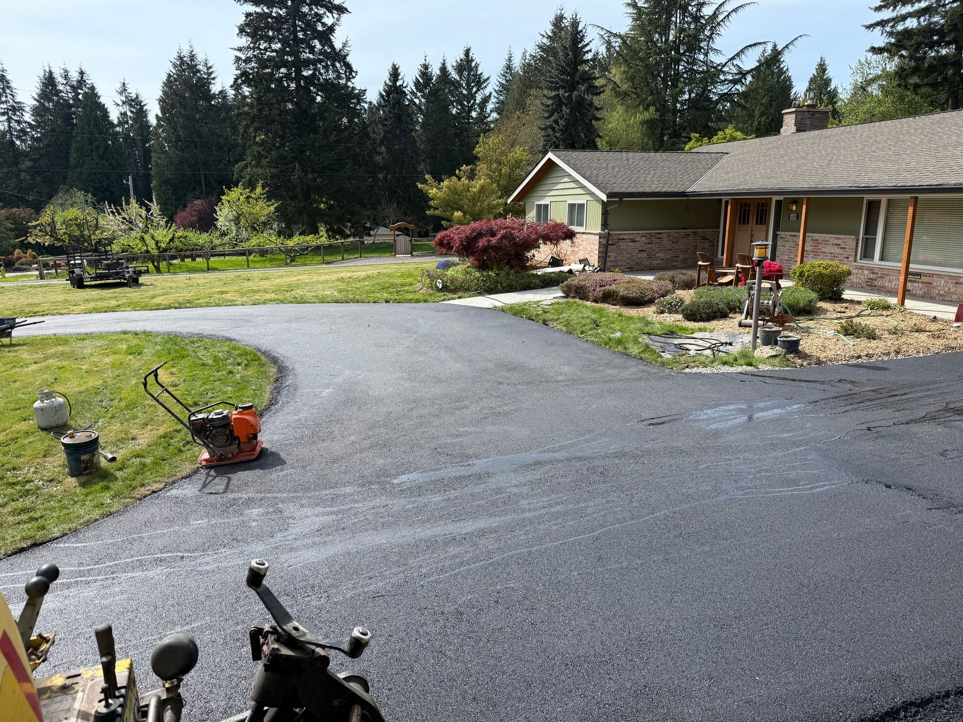 Black paved driveway leading uphill between houses and tall trees on a sunny day.