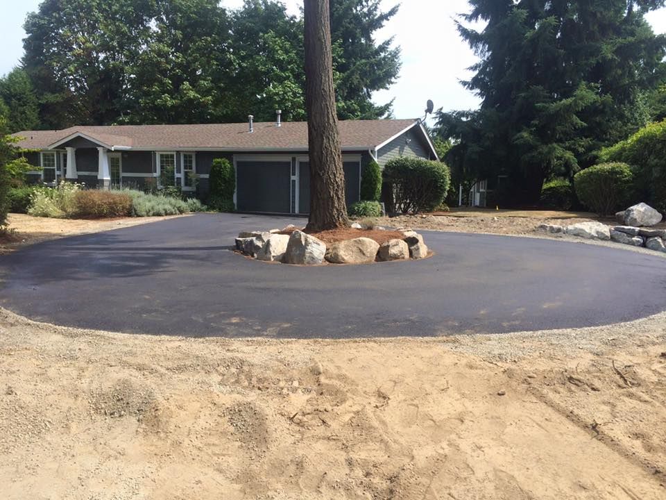 Circular asphalt driveway with a tree in the center, surrounded by rocks, in front of a house.