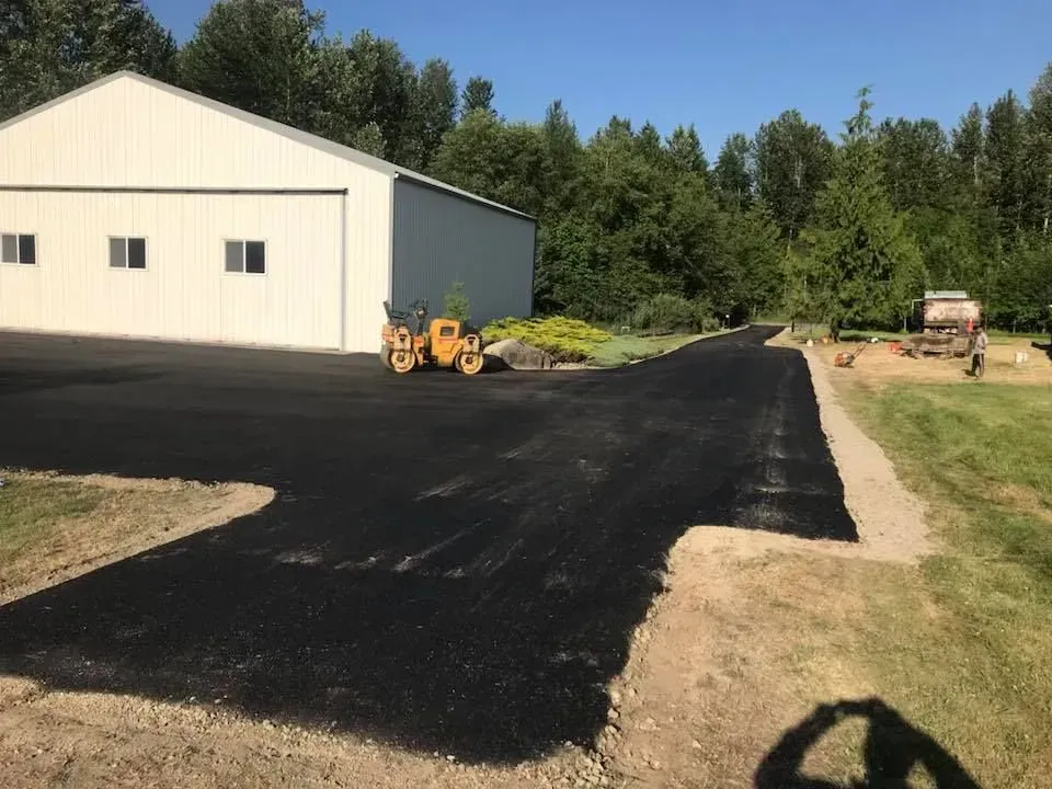 Asphalt driveway under construction next to a white building. A small roller compacts the new surface.
