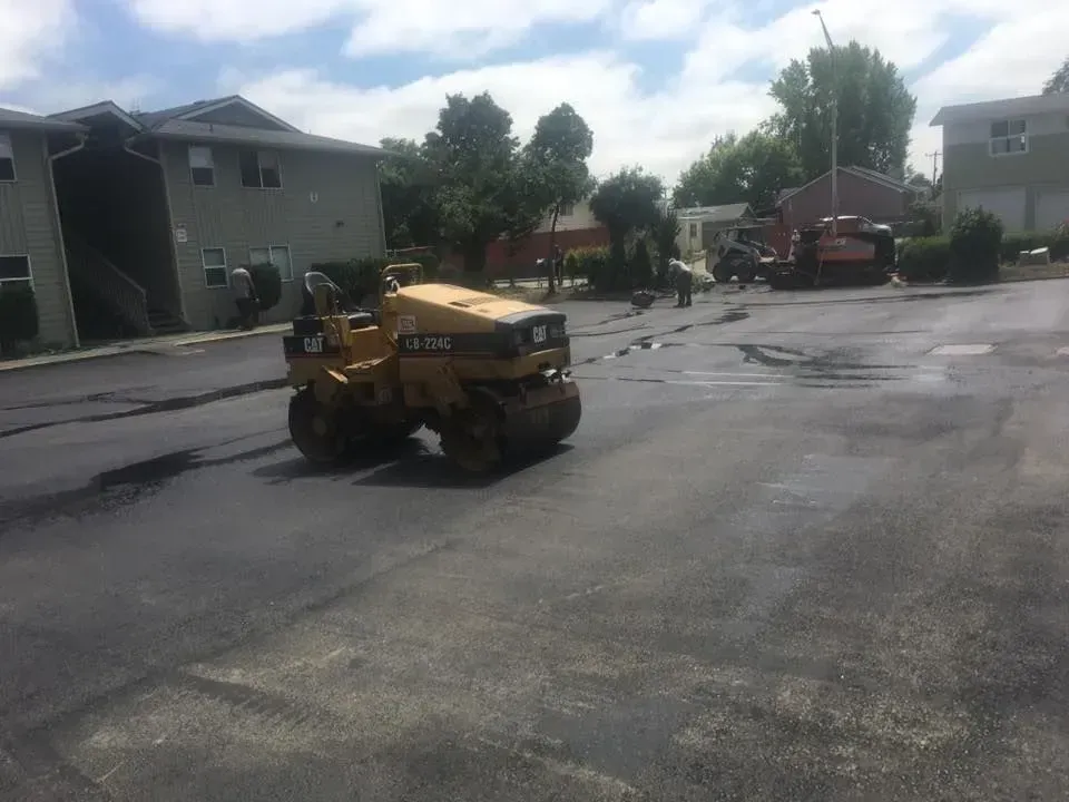 Asphalt roller compacting fresh asphalt on a parking lot, residential buildings in the background.