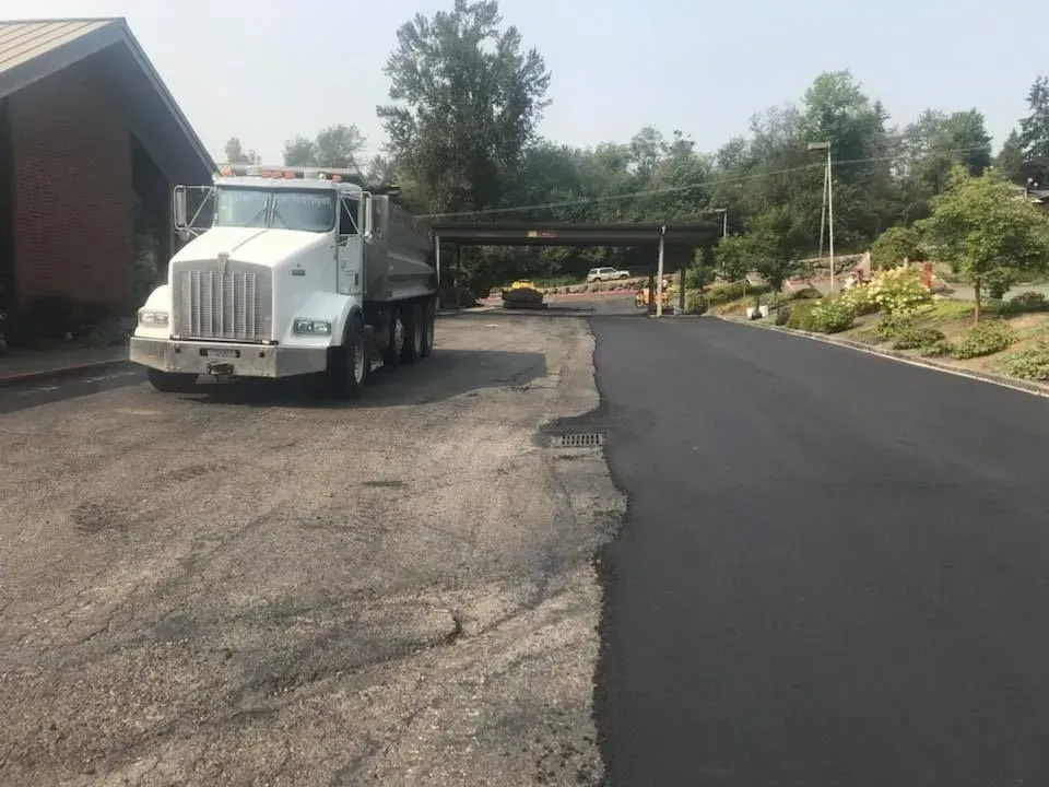 White dump truck next to a freshly paved driveway. A building and trees are in the background.