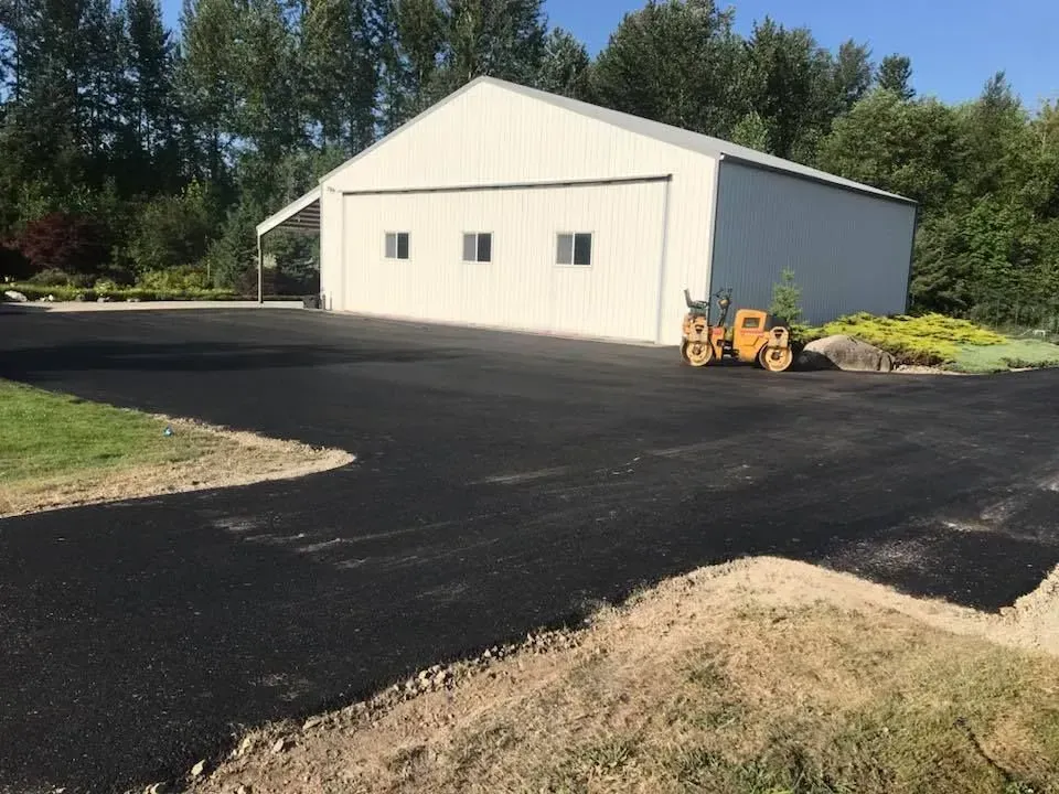 Paved driveway leading to a white metal building with small windows; a small yellow roller sits nearby.