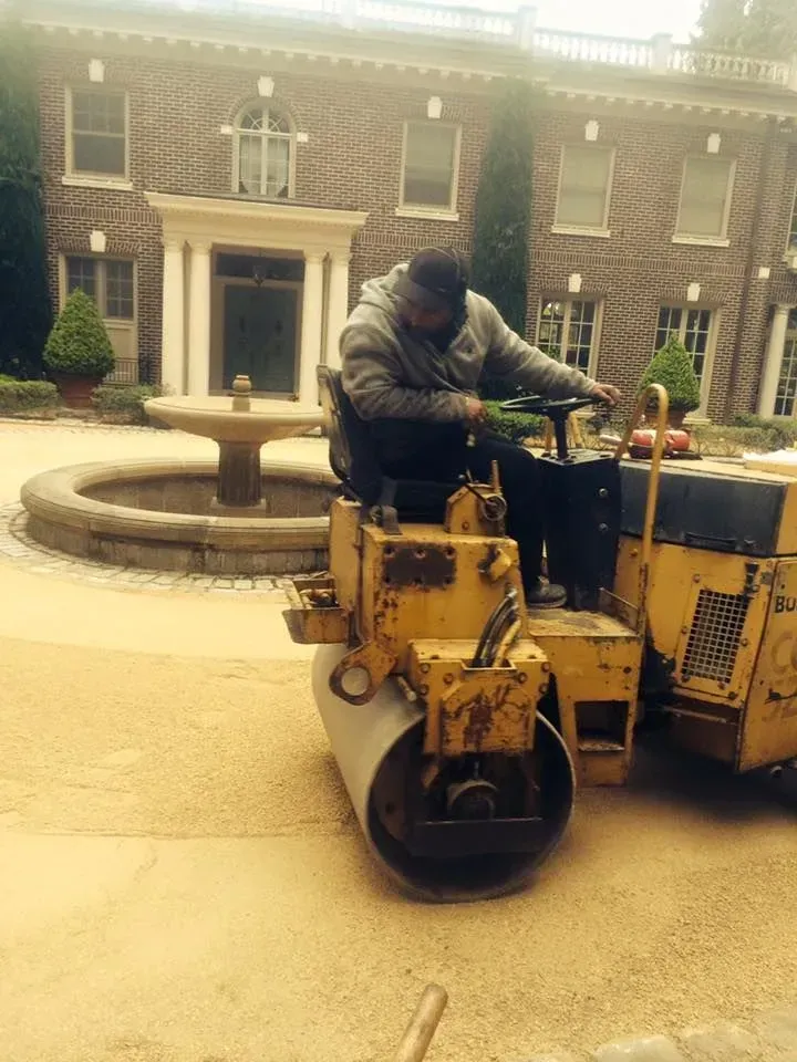 Person operating a yellow road roller in front of a large brick building.