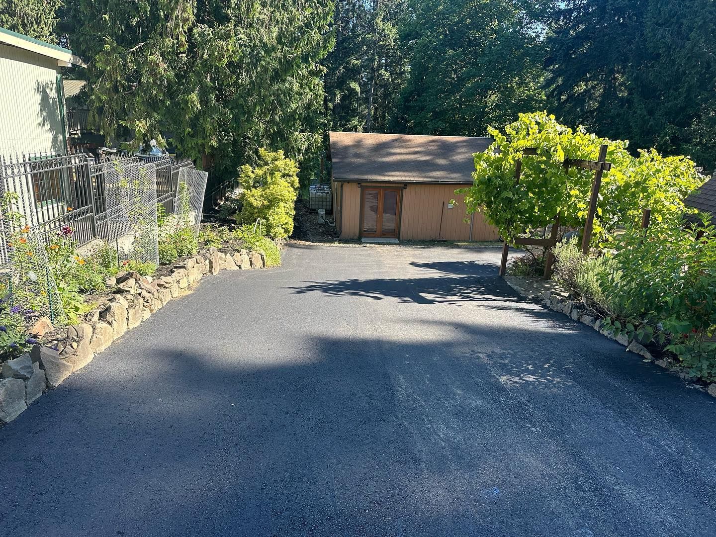 Black asphalt driveway with a shed at the end, bordered by landscaping and a fence.
