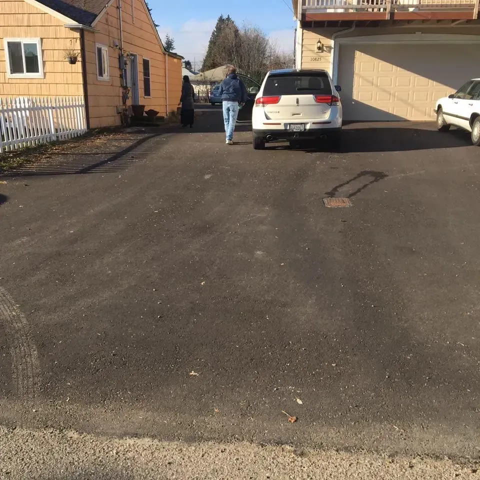 Person walking toward parked white SUV on asphalt driveway. Buildings and fence in background.