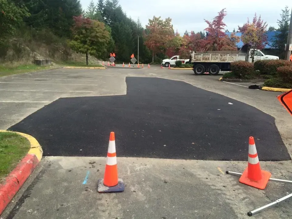 New asphalt patch in a parking lot, orange cones, truck, trees in the background.