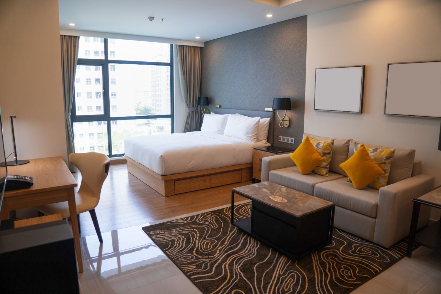 A modern hotel room featuring a queen bed, a seating area with a sofa, two yellow pillows, and a wooden desk.