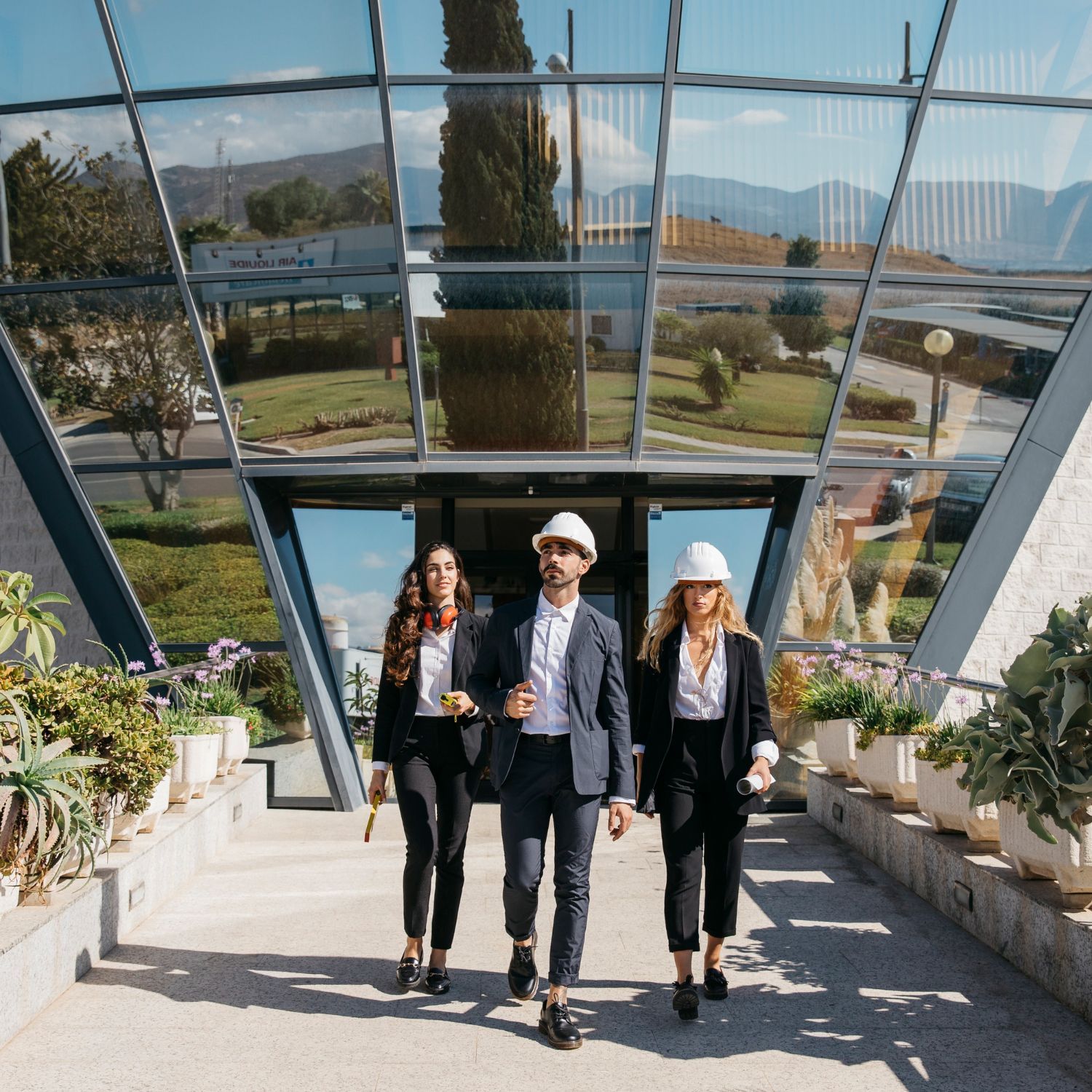 Three professionals in business attire and hard hats walk toward the camera from a modern glass building.