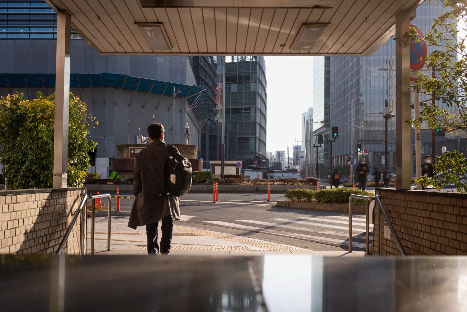 A person with a backpack walks toward a city street from beneath a building entrance overhang on a sunny day.