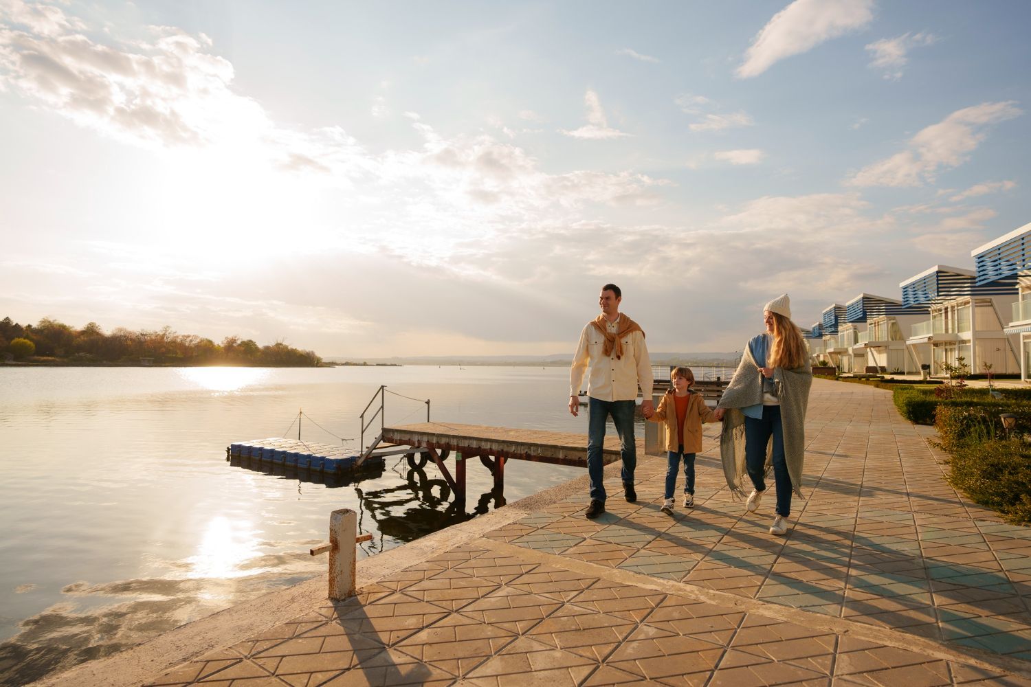 A family walks along a stone pier by a lake during a sunny, golden sunset, with white houses in the background.