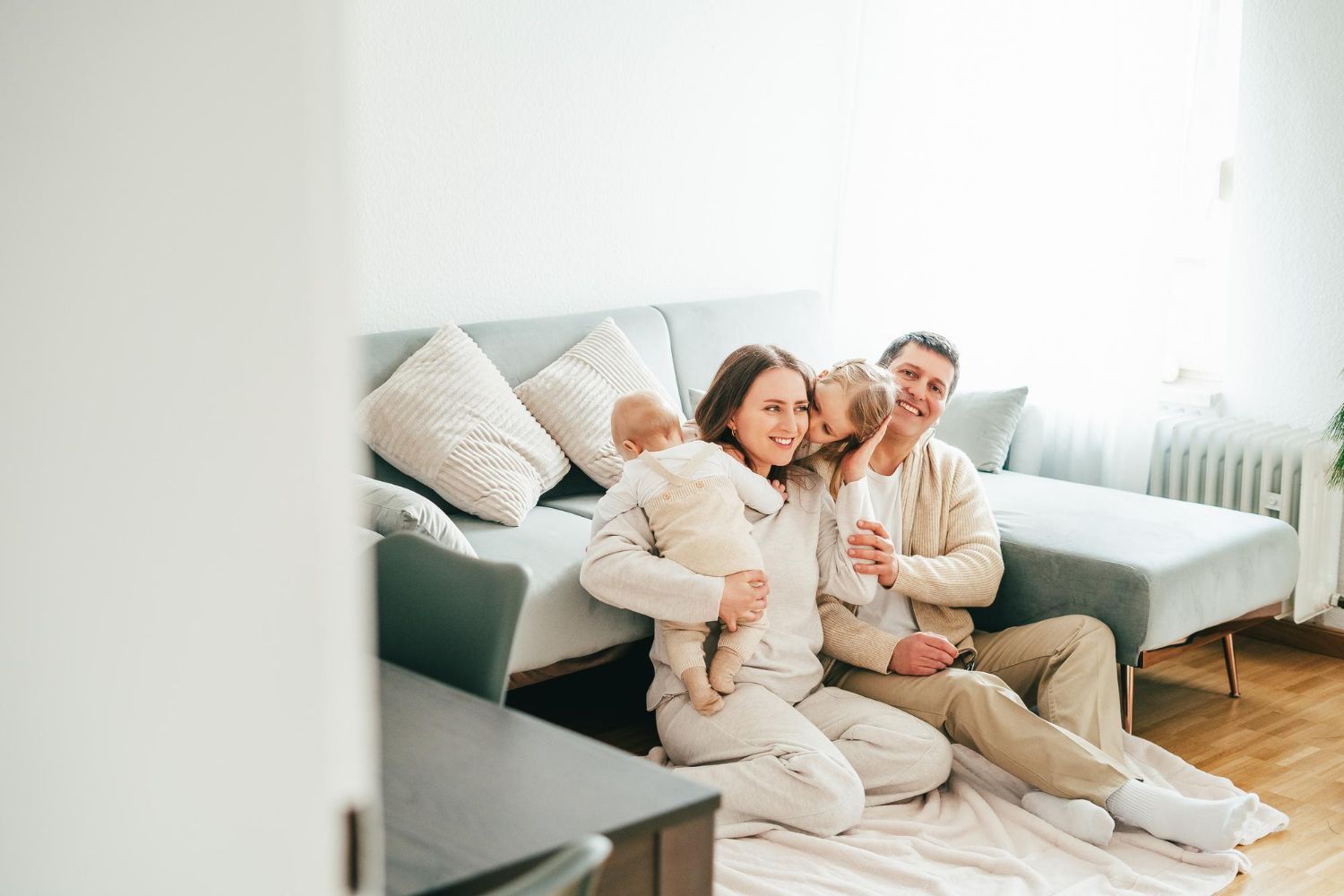 A family of four sits together on a light-colored couch in a brightly lit room, smiling and embracing.