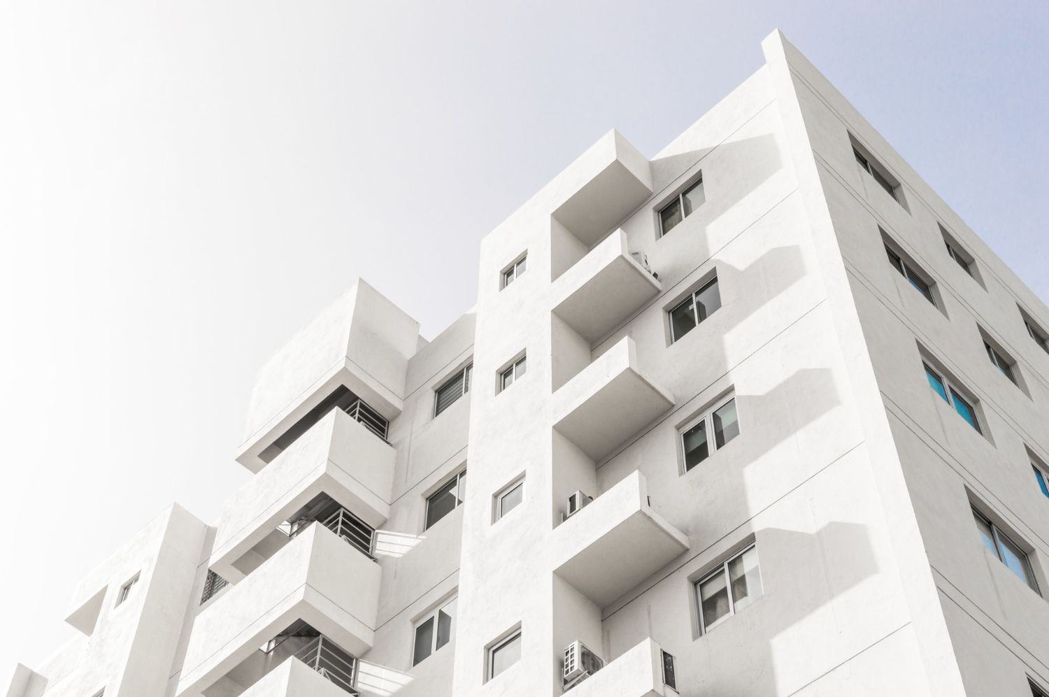 A low-angle view of a white, multi-story modern residential building with projecting balconies under a bright sky.