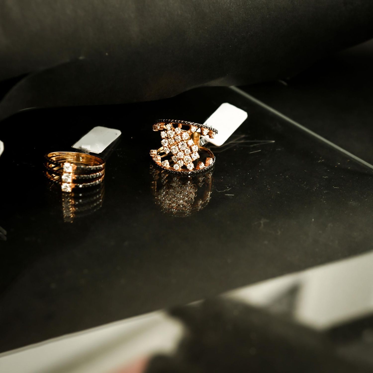Two gold rings with small, sparkling stone accents resting on a dark, reflective surface with price tags attached.