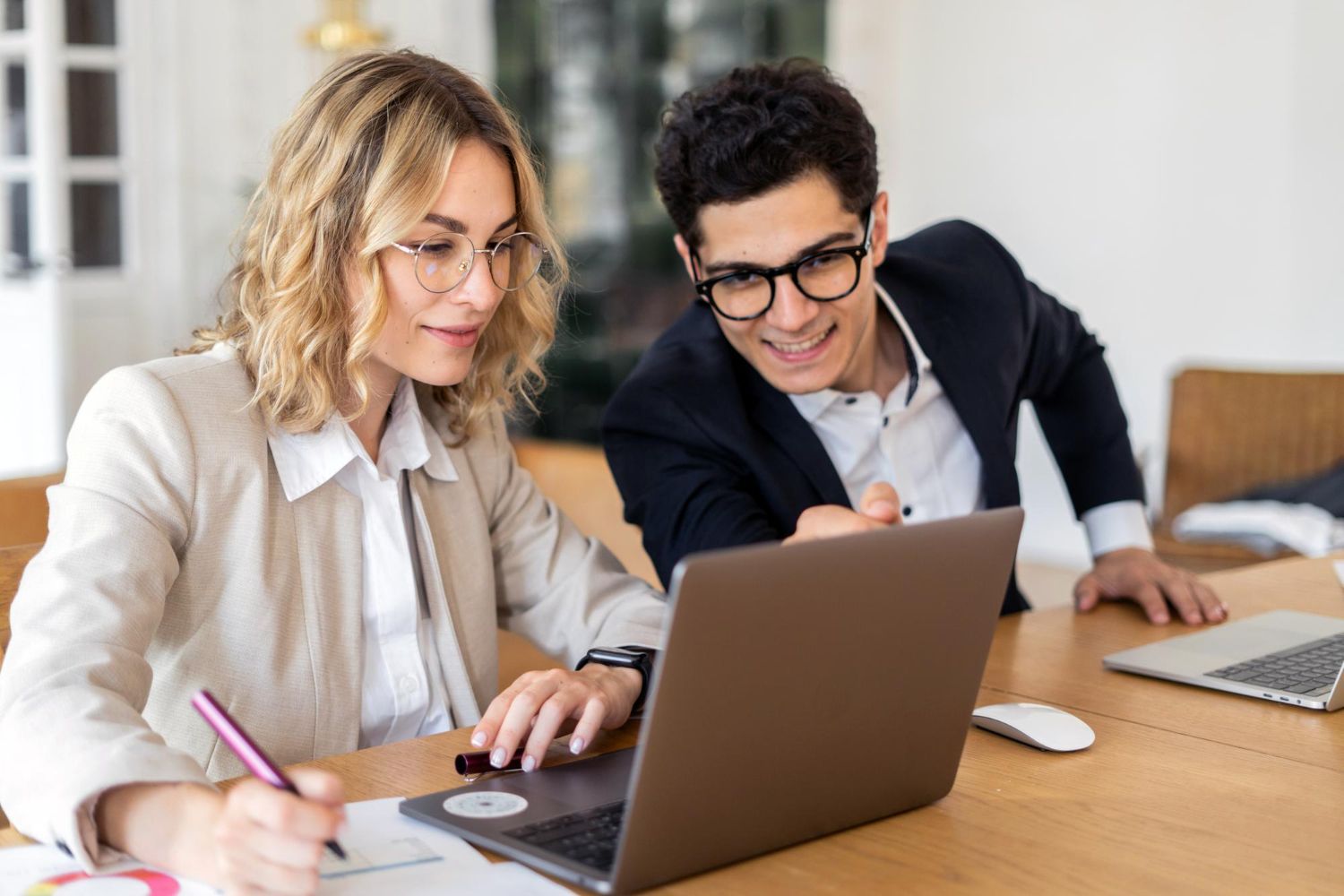 Two people in business attire sit at a desk, looking at a laptop and collaborating while one holds a pen.