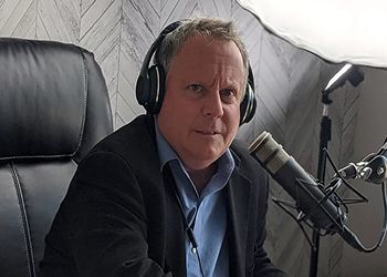 Mr. Hartwell wearing headphones and a blazer sits at a desk with a microphone, facing forward against a patterned wall.