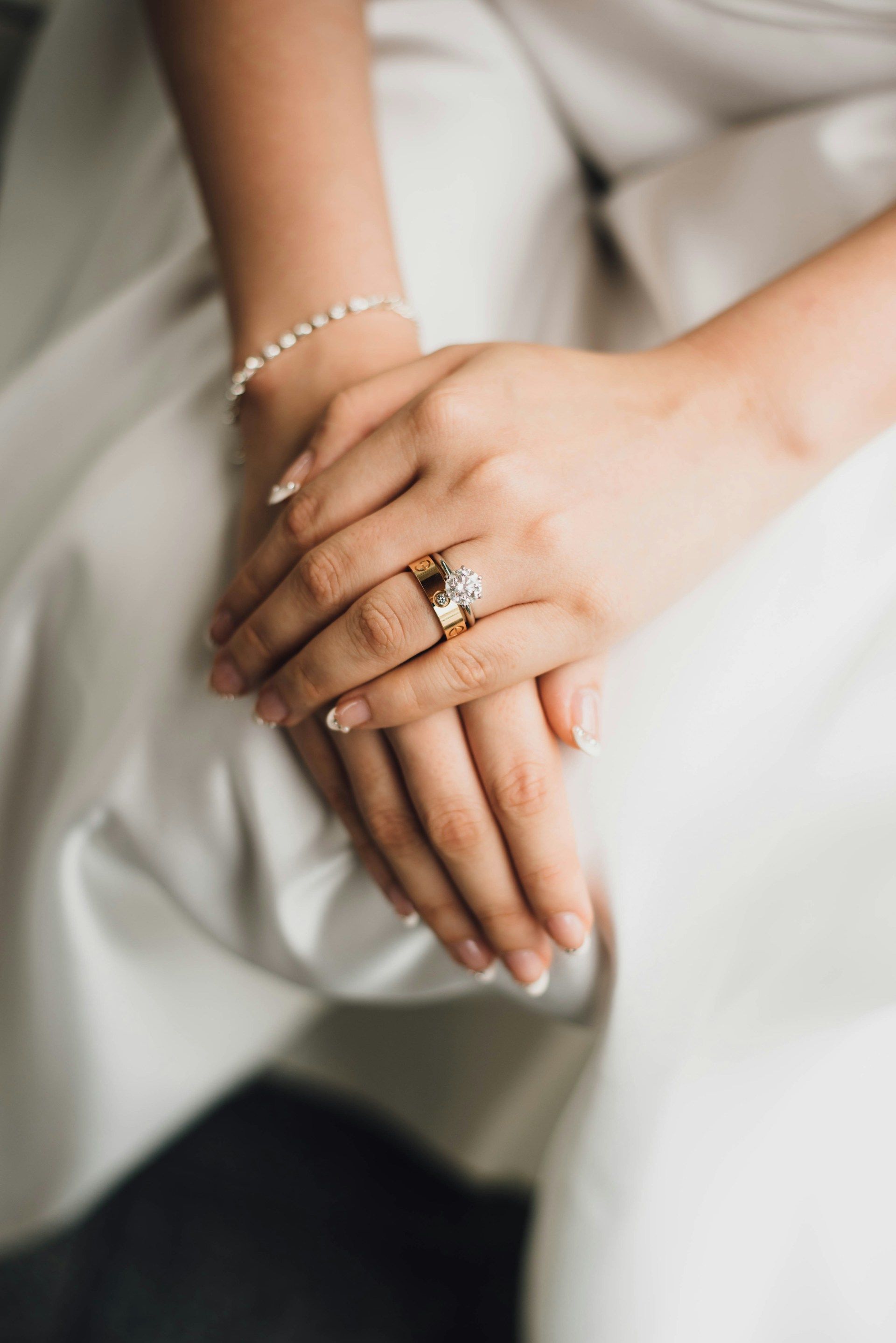 Hands resting on a white fabric, wearing a delicate bracelet and a gold ring with a prominent square-cut stone.