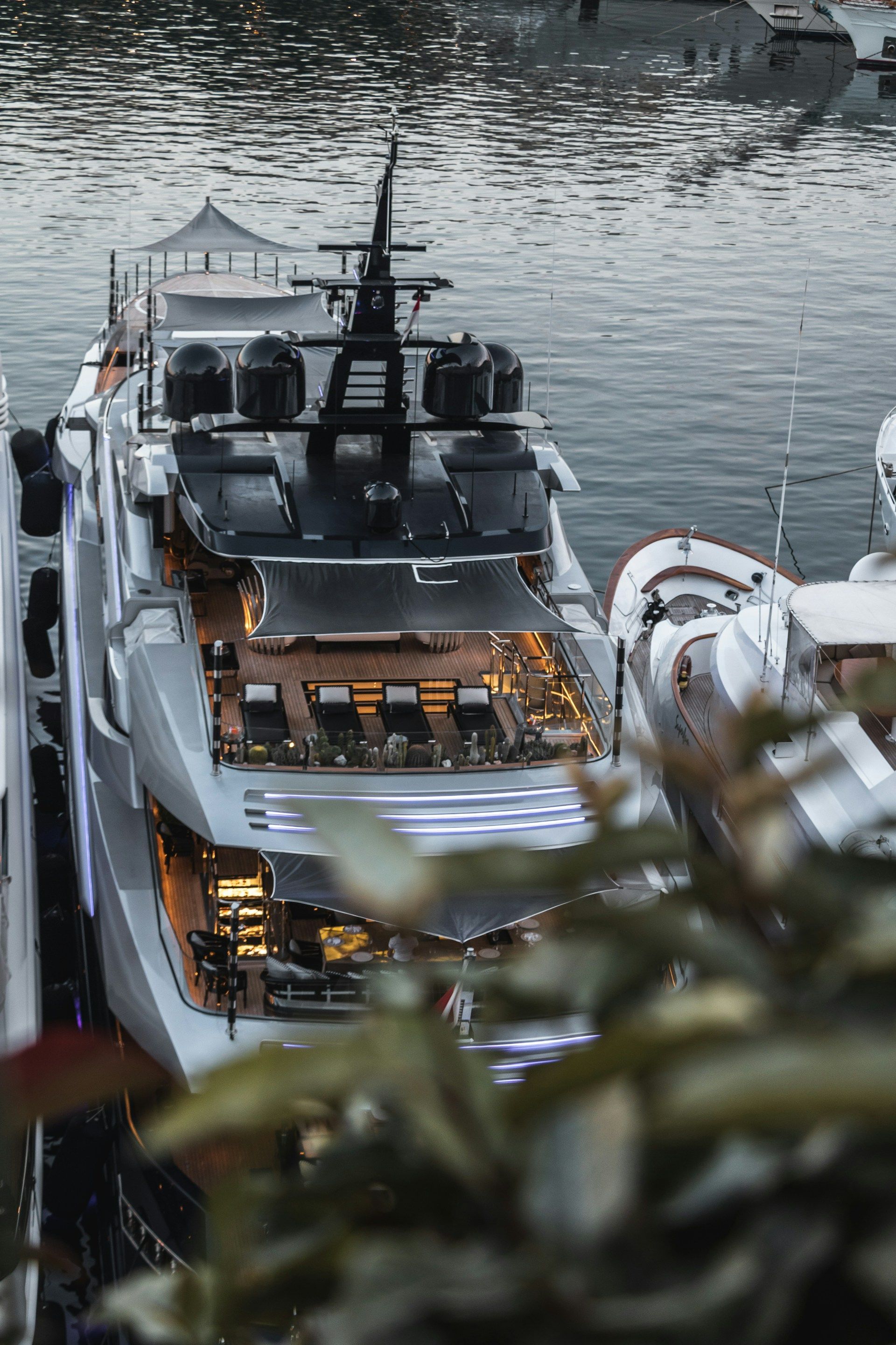 High-angle view of a modern, multi-level luxury yacht docked in water, partially obscured by foreground foliage.