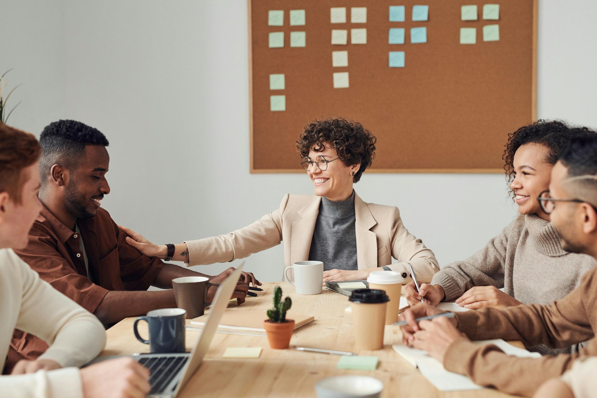 A team sits around a wooden table in an office meeting, with a woman gesturing as she speaks to colleagues.
