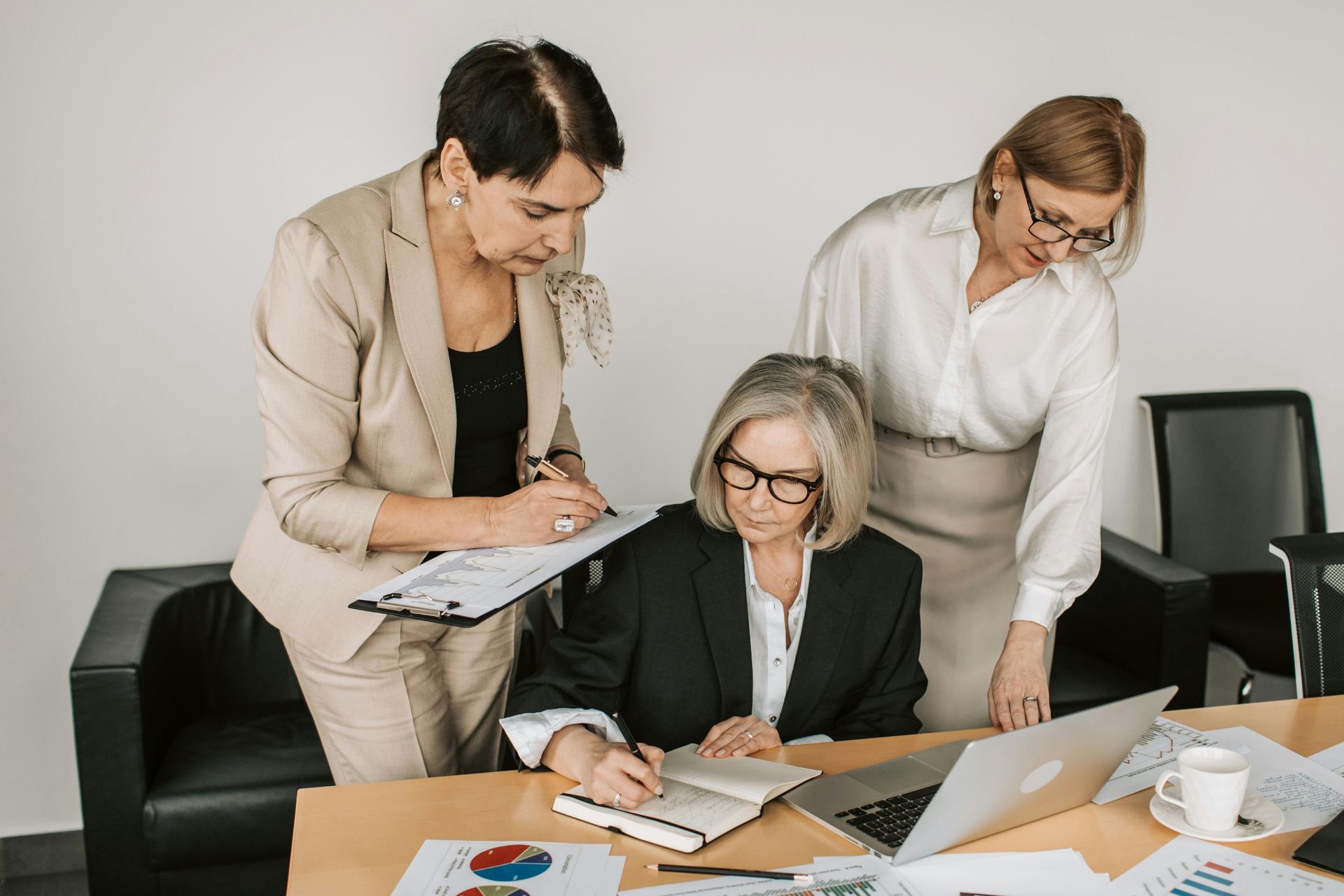Three professionals in business attire collaborate at a table with laptops, documents, and charts.