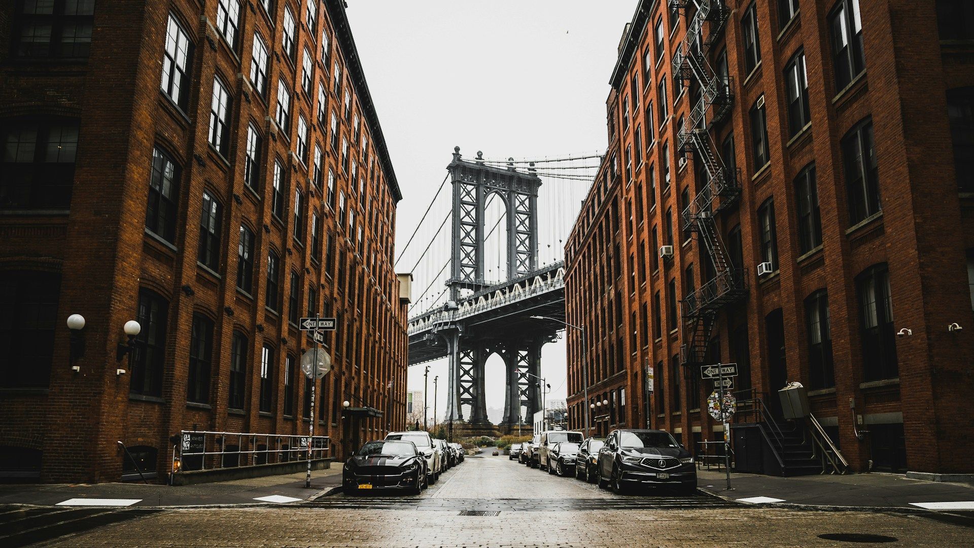 The Manhattan Bridge framed by two rows of red brick industrial buildings on a cobblestone street in New York City.