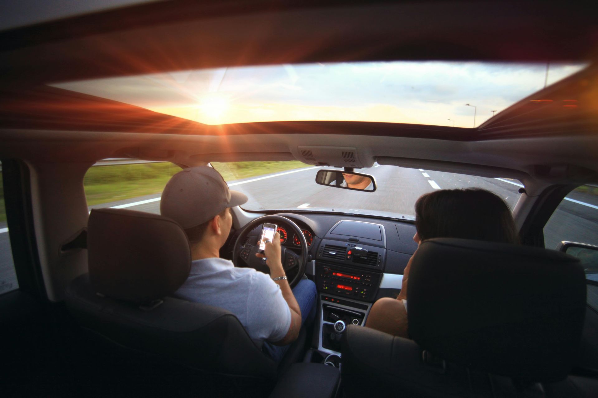A driver uses a smartphone while operating a car on a road at sunset, with a passenger beside them.
