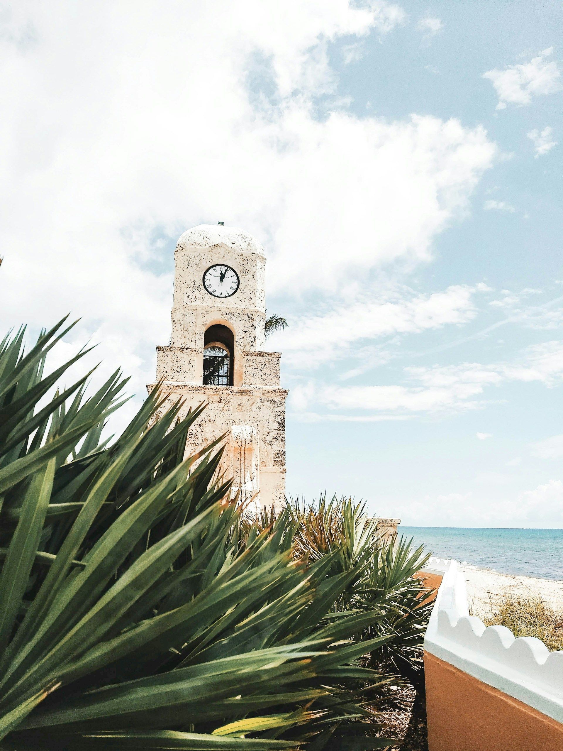 A stone clock tower stands near a sandy beach and ocean under a bright, cloudy sky, framed by lush tropical plants.