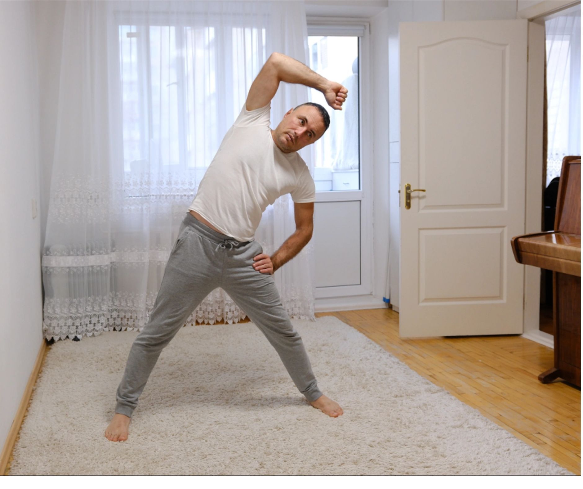 Man stretching torso in a home setting, with arm overhead and hand on hip.