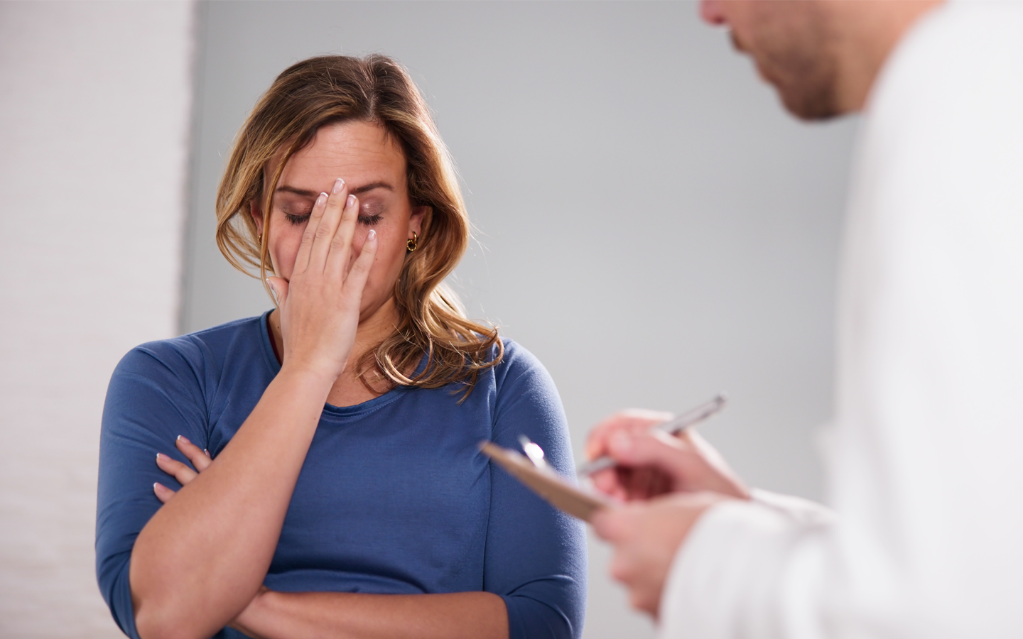 Woman with hand on face, appears distressed, speaking to doctor with clipboard in a consultation room.