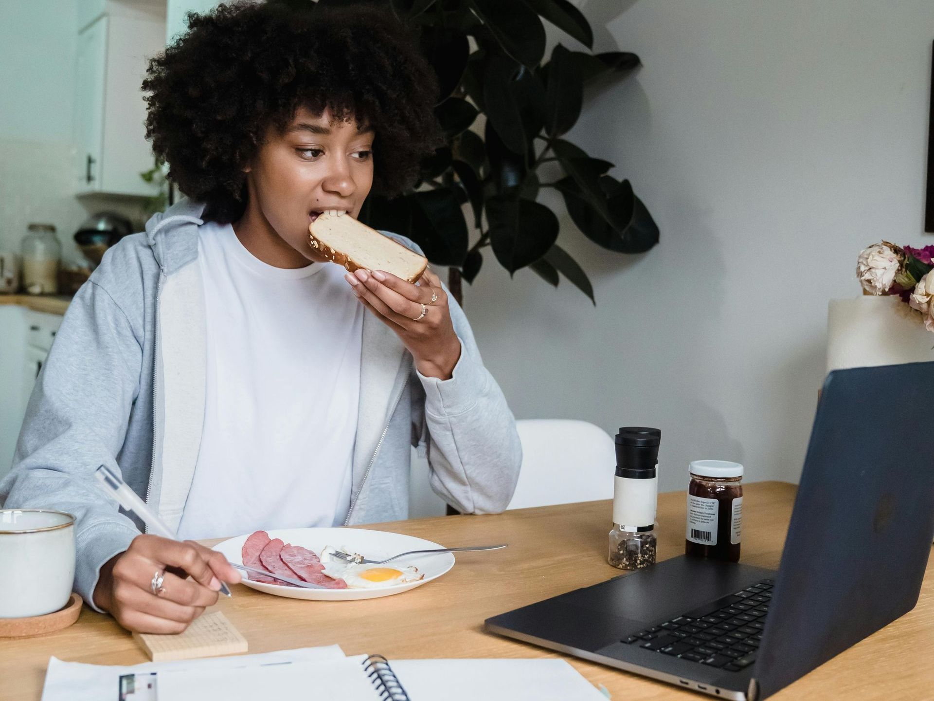 A woman is sitting at a table eating a sandwich and looking at a laptop.