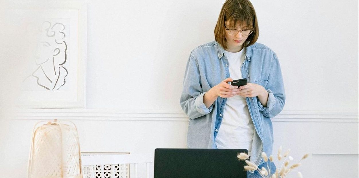 A woman is standing in front of a laptop computer looking at her phone.