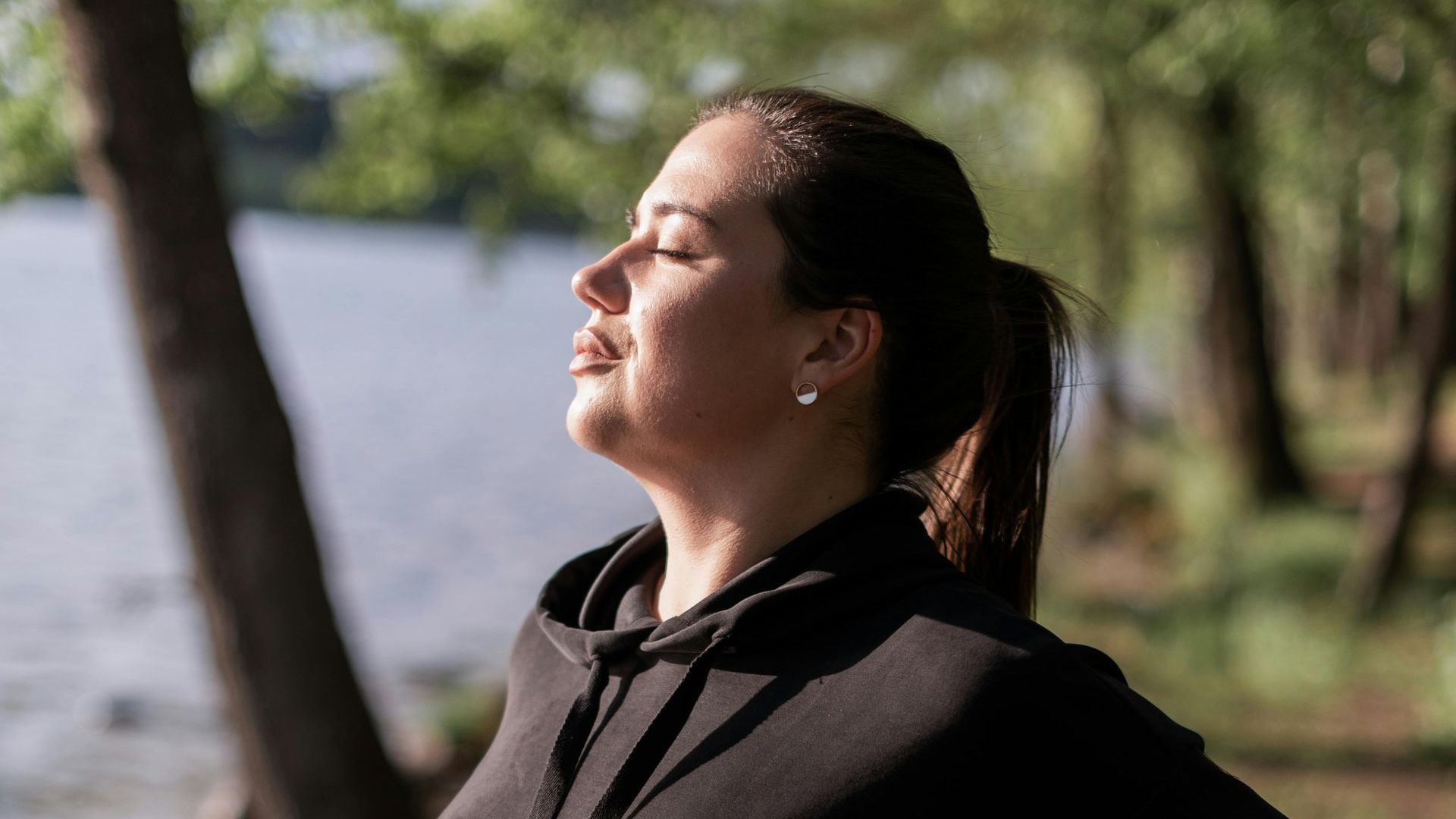 A woman is standing in front of a lake with her eyes closed.