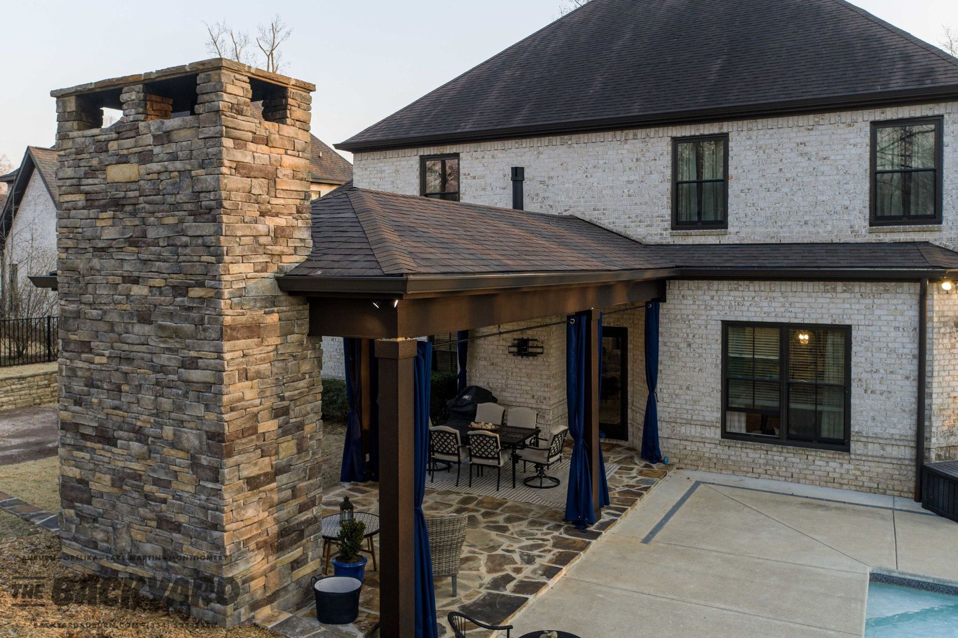 Stone chimney next to a white brick house with a covered patio and swimming pool.