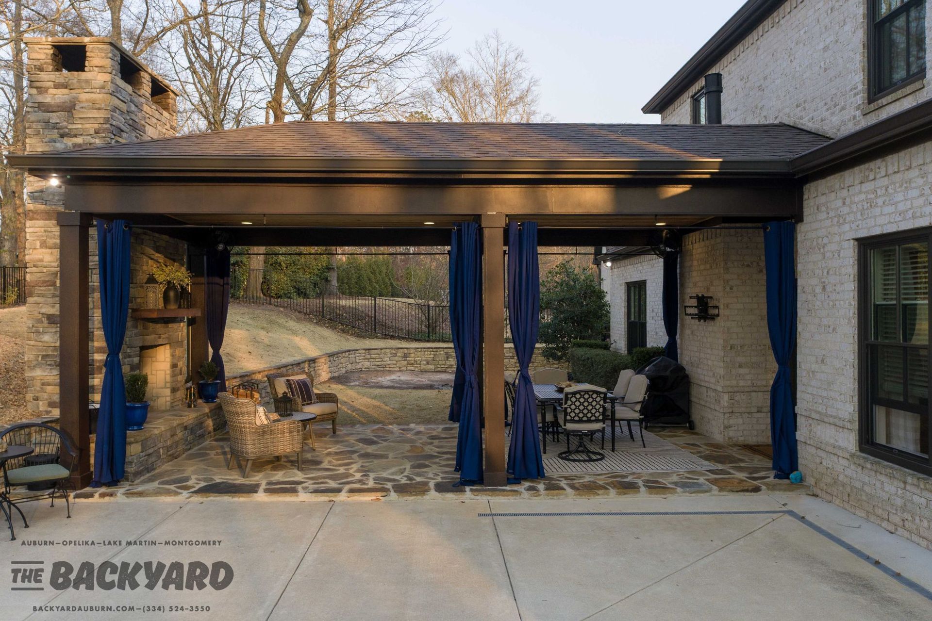 Covered patio with stone fireplace, blue curtains, and outdoor seating. Brick house in the background.