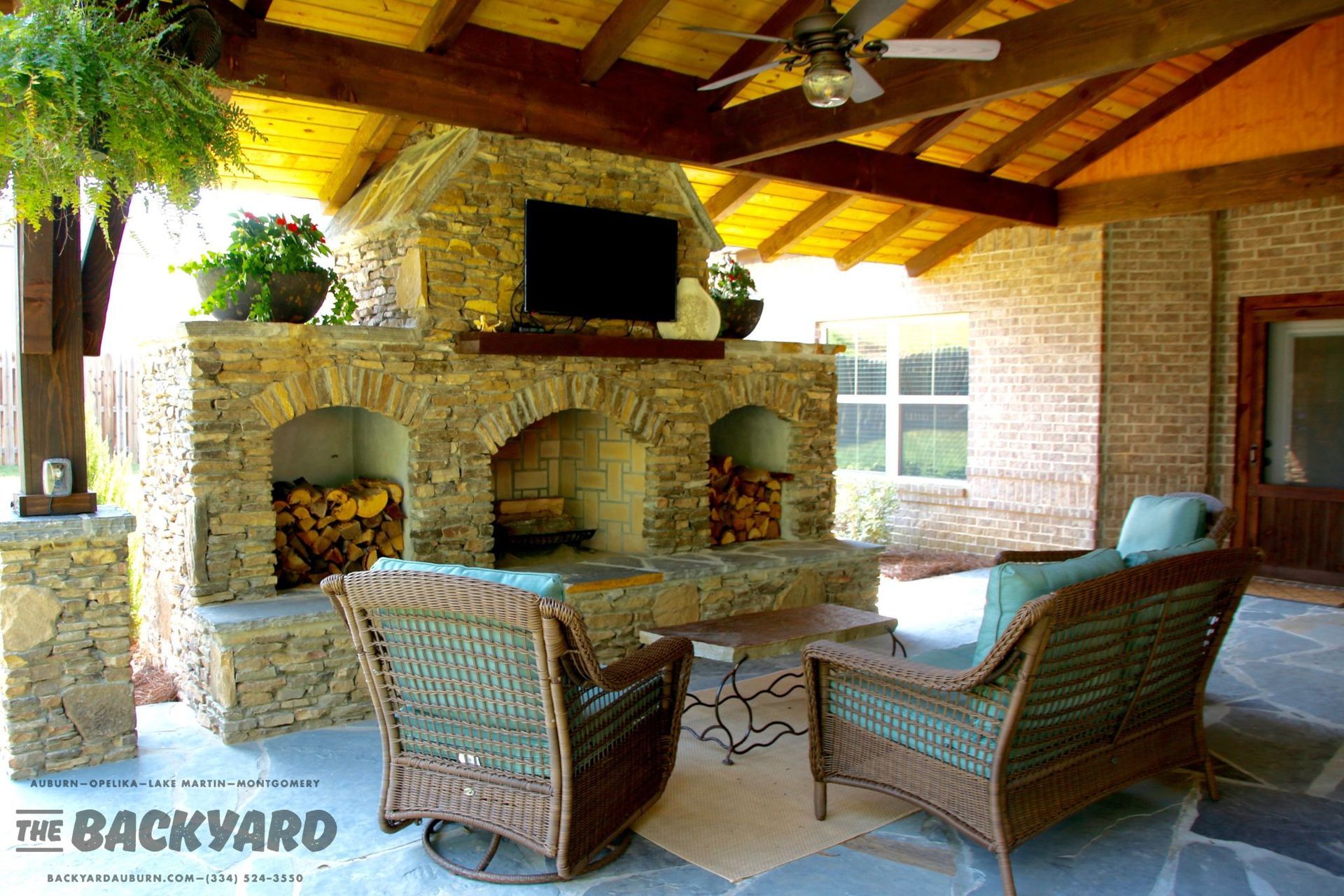 Stone outdoor fireplace with firewood, TV, seating, and wood ceiling.