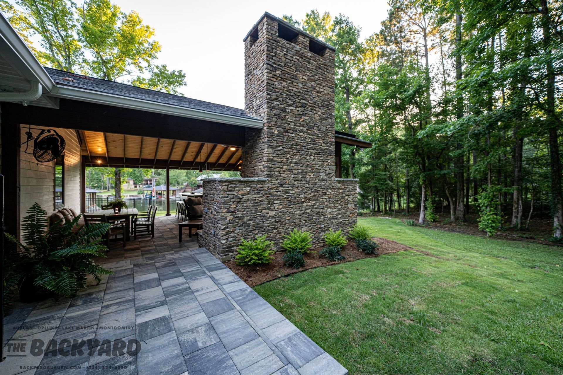Outdoor patio with stone chimney and paved walkway, overlooking trees and lawn.